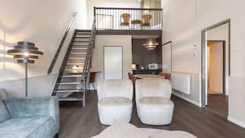 Un vaste espace de vie comprenant un escalier menant à une mezzanine, un canapé, des fauteuils et un coin repas avec une table et des chaises.