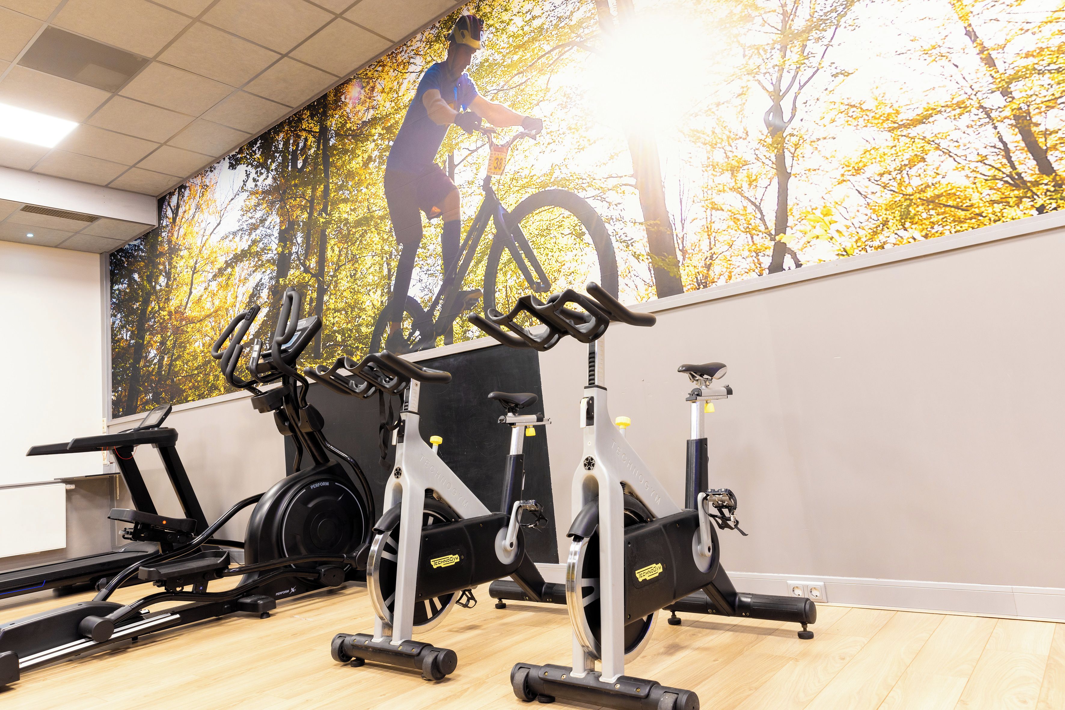 Ein Fitnessstudio mit Ergometer, Laufbändern und einer Wandmalerei eines Radfahrers.