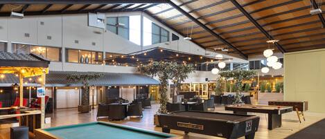 Un vaste espace de loisirs intérieur comprenant des tables de billard, des sièges et un haut plafond avec des poutres apparentes.
