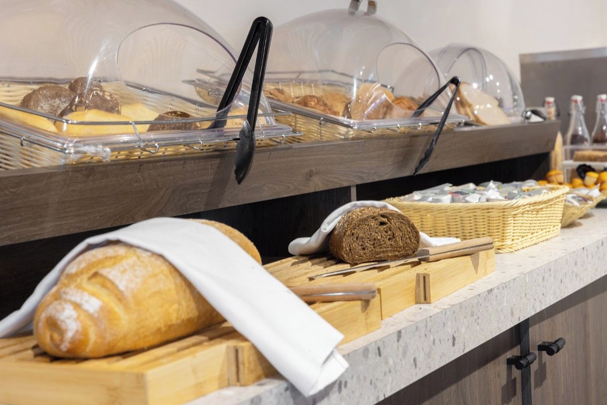 Disposição de buffet com diversos alimentos sob redomes de vidro, uma barra de pão em tábua de corte e uma cesta de vime.