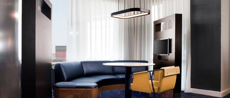 A modern hotel room with a curved black leather sofa, a round wooden table, a yellow chair, a flat-screen TV mounted on the wall, and a large window with sheer curtains.