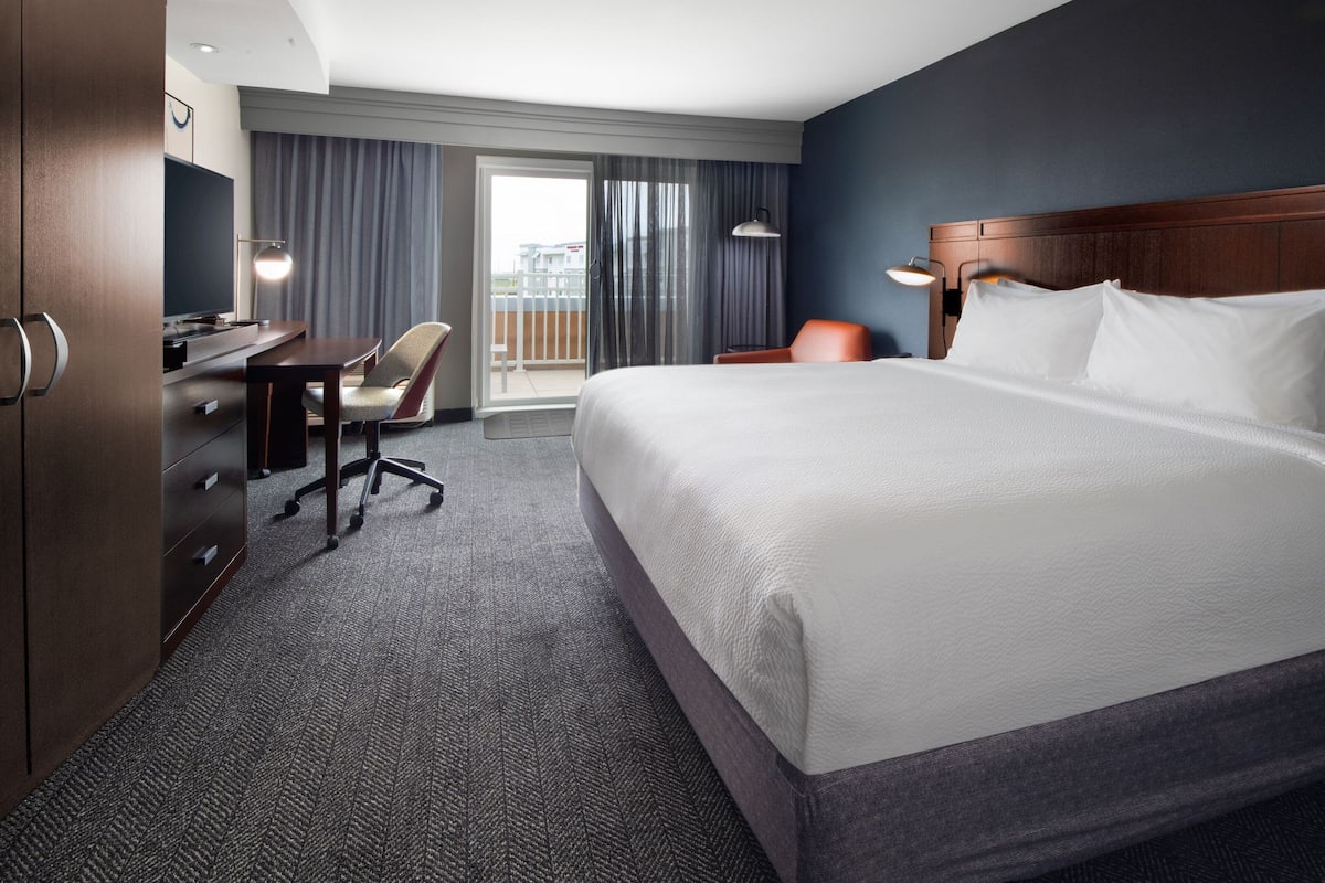 A hotel room with a large bed, a desk with a chair, a television, and a view of the outdoors through a sliding door.