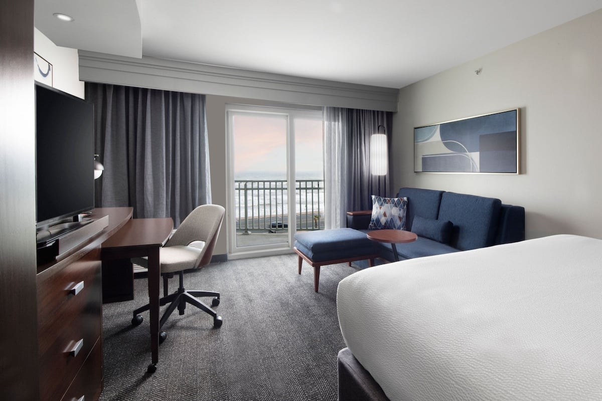A hotel room with a bed, a desk, a chair, a small table, and a view of the outdoors through a large window.