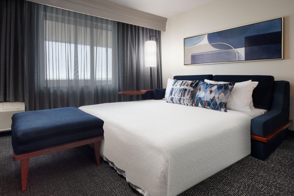 A hotel room with a large bed, a bench, a bedside table, a lamp, and a painting on the wall.