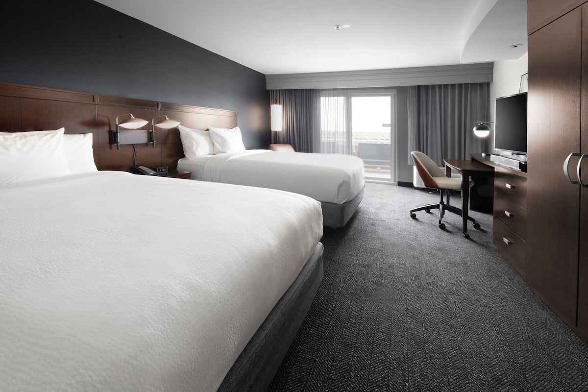 A hotel room with two beds, a desk, a chair, a TV, and a large window with curtains.