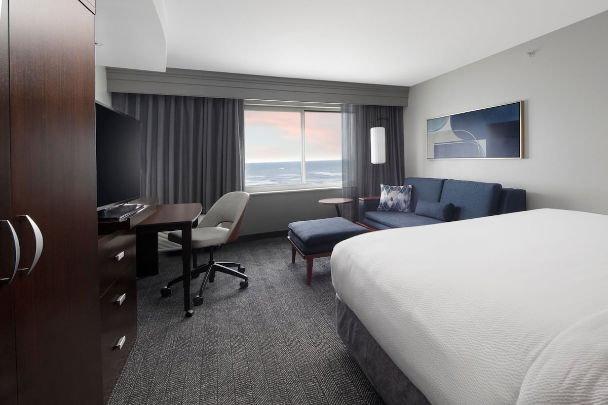 A hotel room with a bed, a desk, a chair, a small table, and a window with a view of the ocean.