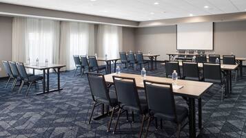 A conference room with multiple tables and chairs, equipped with water bottles, a projector screen, and a large window with curtains.