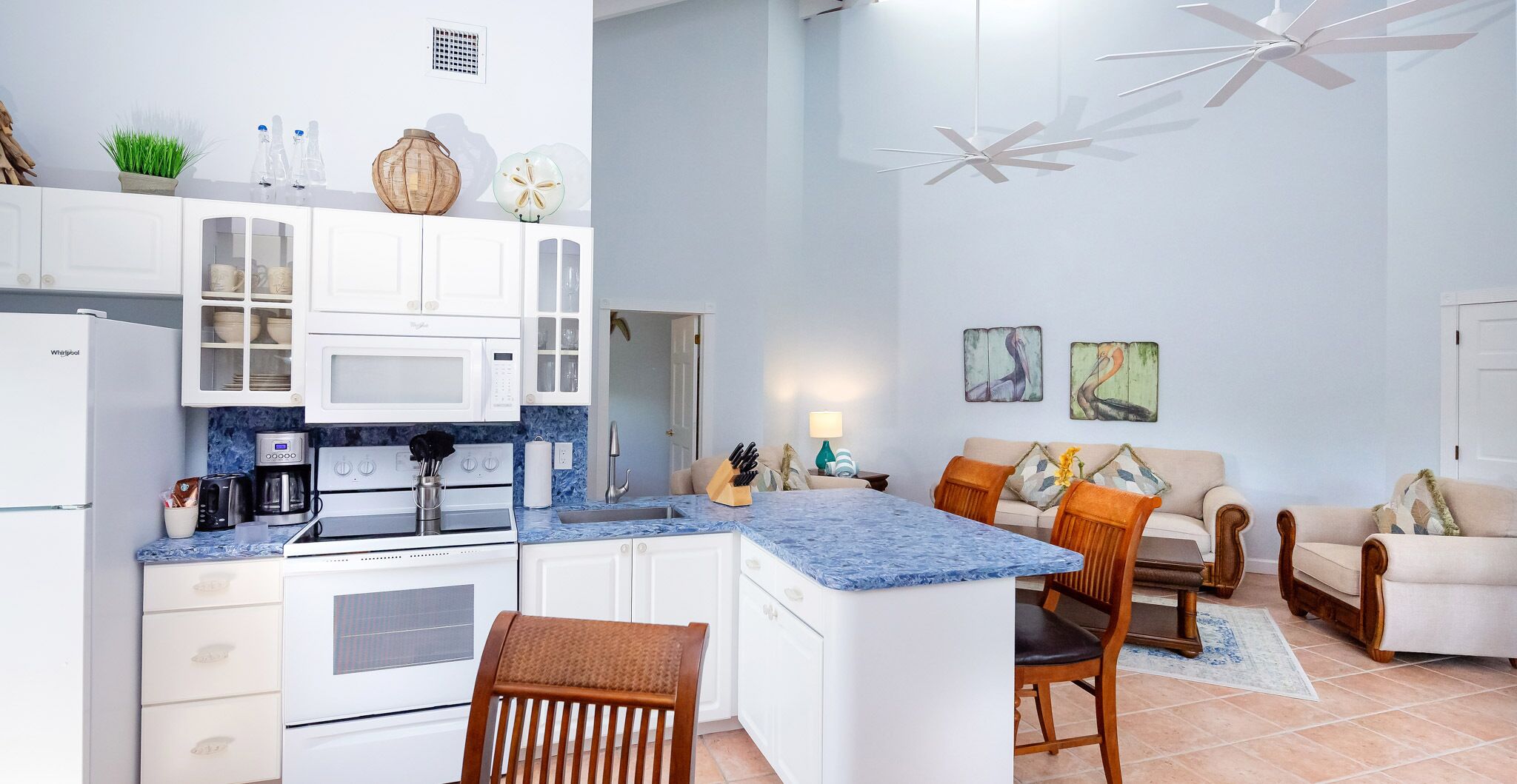 A modern kitchen with white cabinets, a built-in microwave, and a granite countertop. There is a dining area with a wooden table and chairs, and a living room with a sofa and a coffee table.