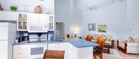 A modern kitchen with white cabinets, a built-in microwave, and a granite countertop. There is a dining area with a wooden table and chairs, and a living room with a sofa and a coffee table.