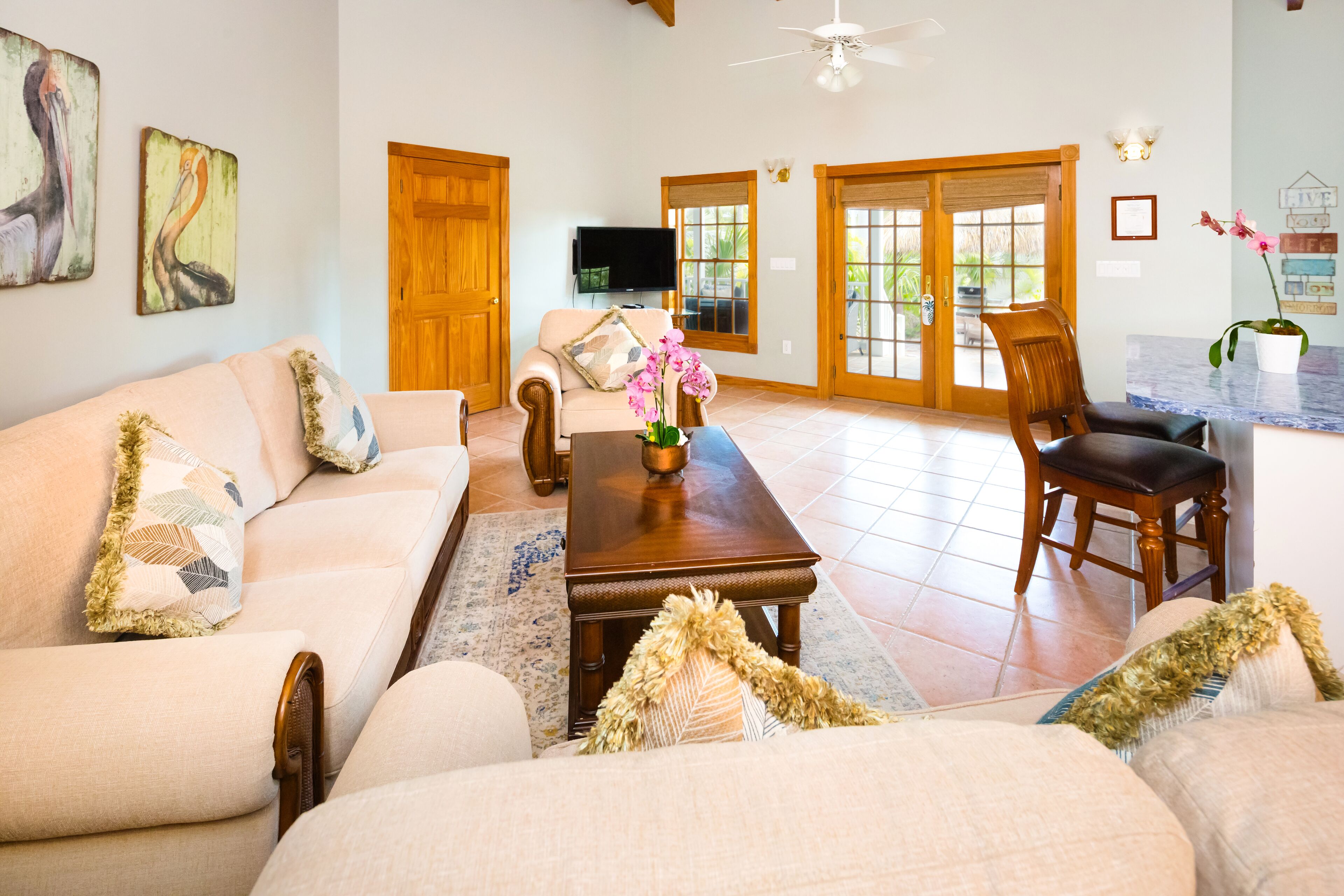 A spacious living room with a beige sofa, a wooden coffee table, and a television. There are wooden doors and a large window with a view of greenery.