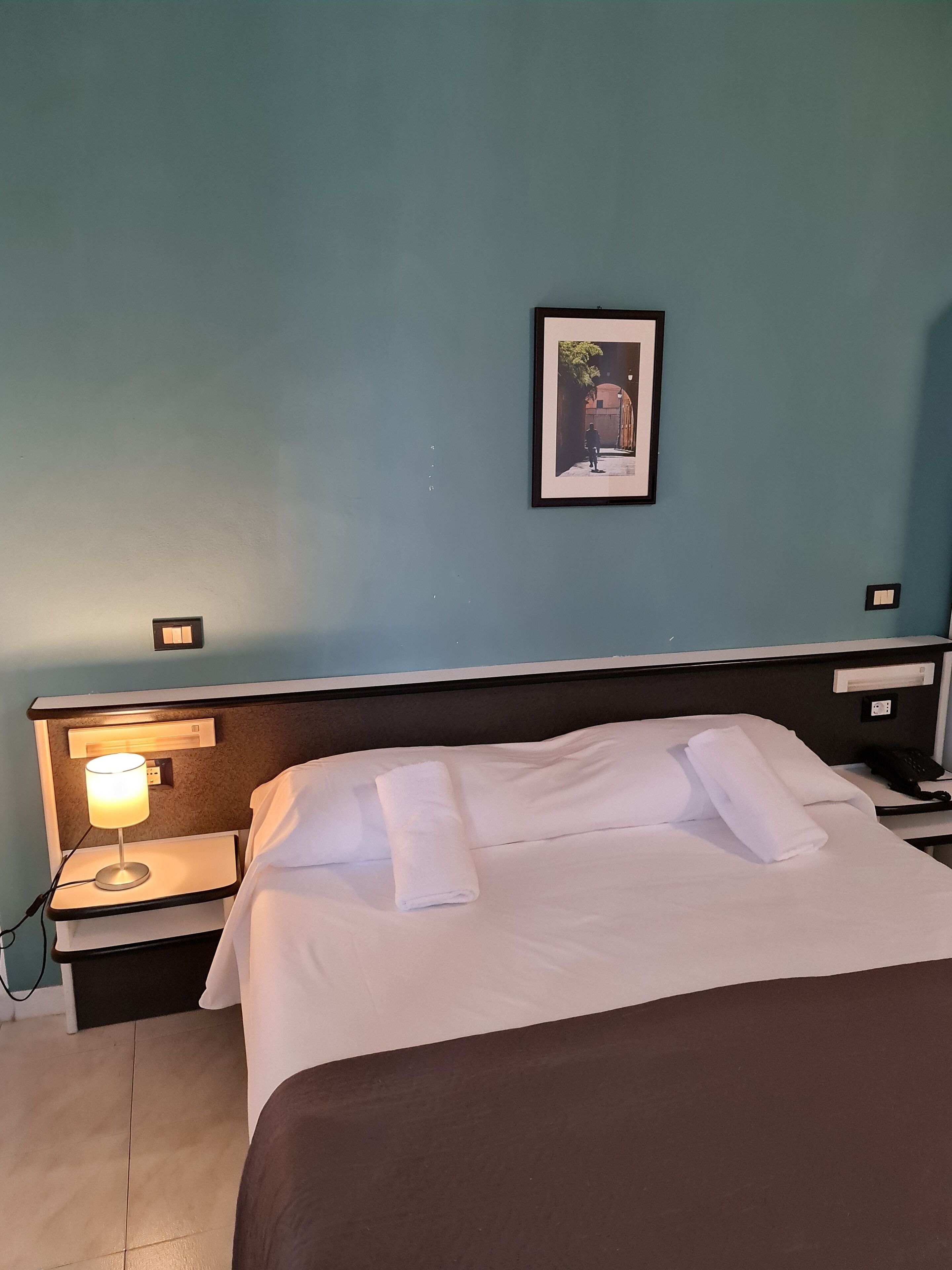 Habitación de hotel con una cama, una mesita de noche con lámpara, un cuadro en la pared y un teléfono sobre la mesita.