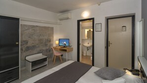 Habitación de hotel compacta con cama, escritorio con computadora, silla, mesita y puerta que da a un baño.