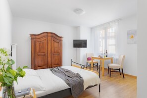 Ein Schlafzimmer mit Bett, Holzschrank, Fernseher, Esstisch, Stühlen und einem Fenster mit Vorhängen.