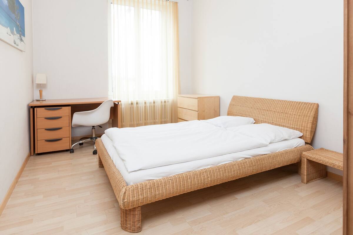 A bedroom with a wooden bed, a desk with a chair, a nightstand, and a window with sheer curtains.