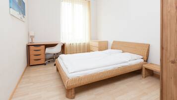 Ein Schlafzimmer mit einem hölzernen Bett, einem Schreibtisch mit Stuhl, einem Nachttisch und einem Fenster mit durchsichtigen Vorhängen.