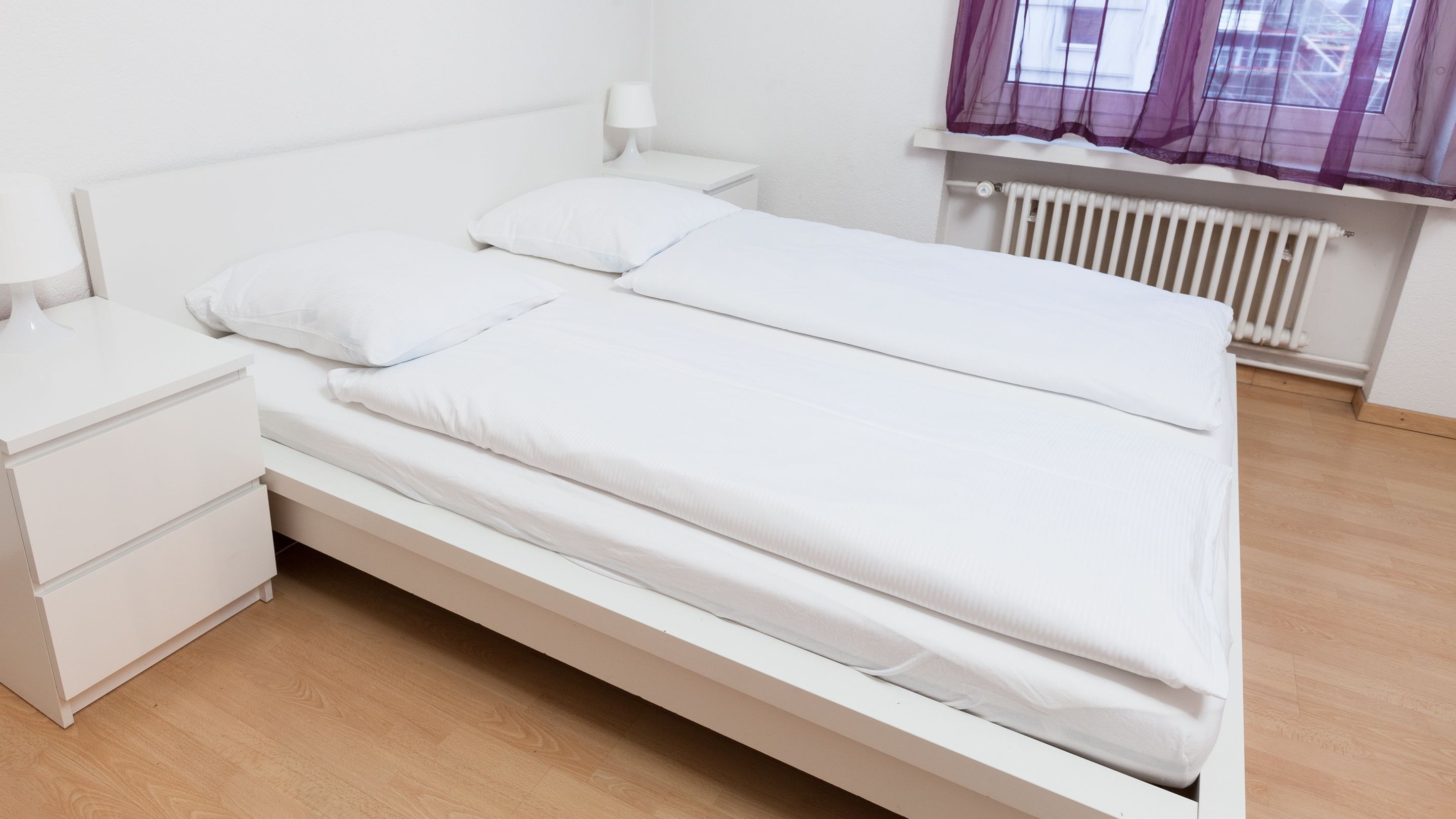 A single-bed room with a white bed, a nightstand, a lamp, and a window with purple curtains.