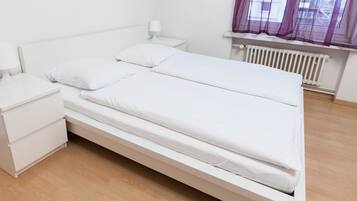 A single-bed room with a white bed, a nightstand, a lamp, and a window with purple curtains.