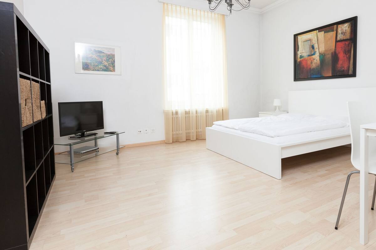 A modern, minimalistic bedroom with a bed, a TV on a glass stand, a desk, and a painting on the wall.