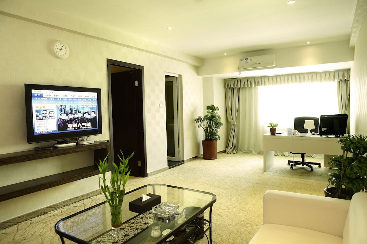 A hotel room with a TV, a glass coffee table, a sofa, a desk with a computer, and a potted plant.