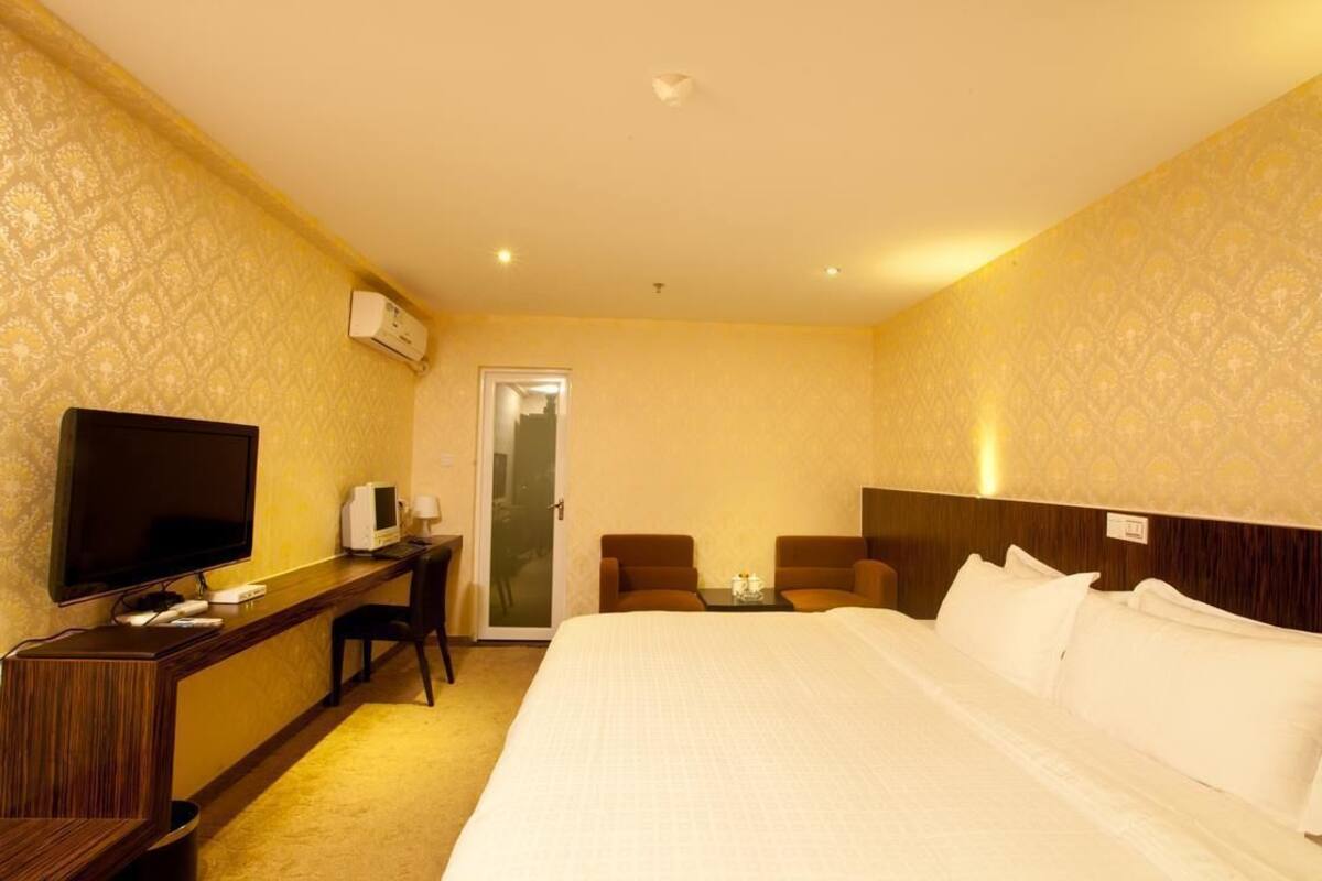 A hotel room with a bed, a desk with a TV, a chair, and a small table with a candle.