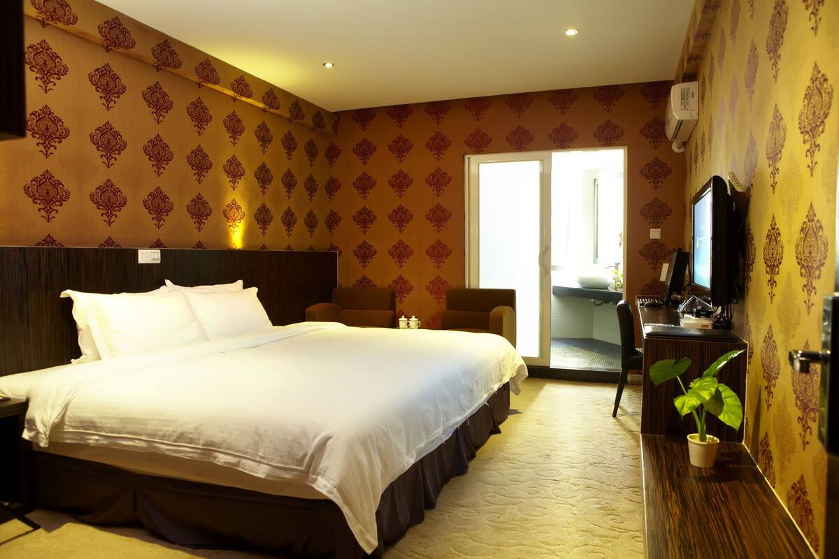 A hotel room with a large bed, a desk with a computer, a chair, and a potted plant.