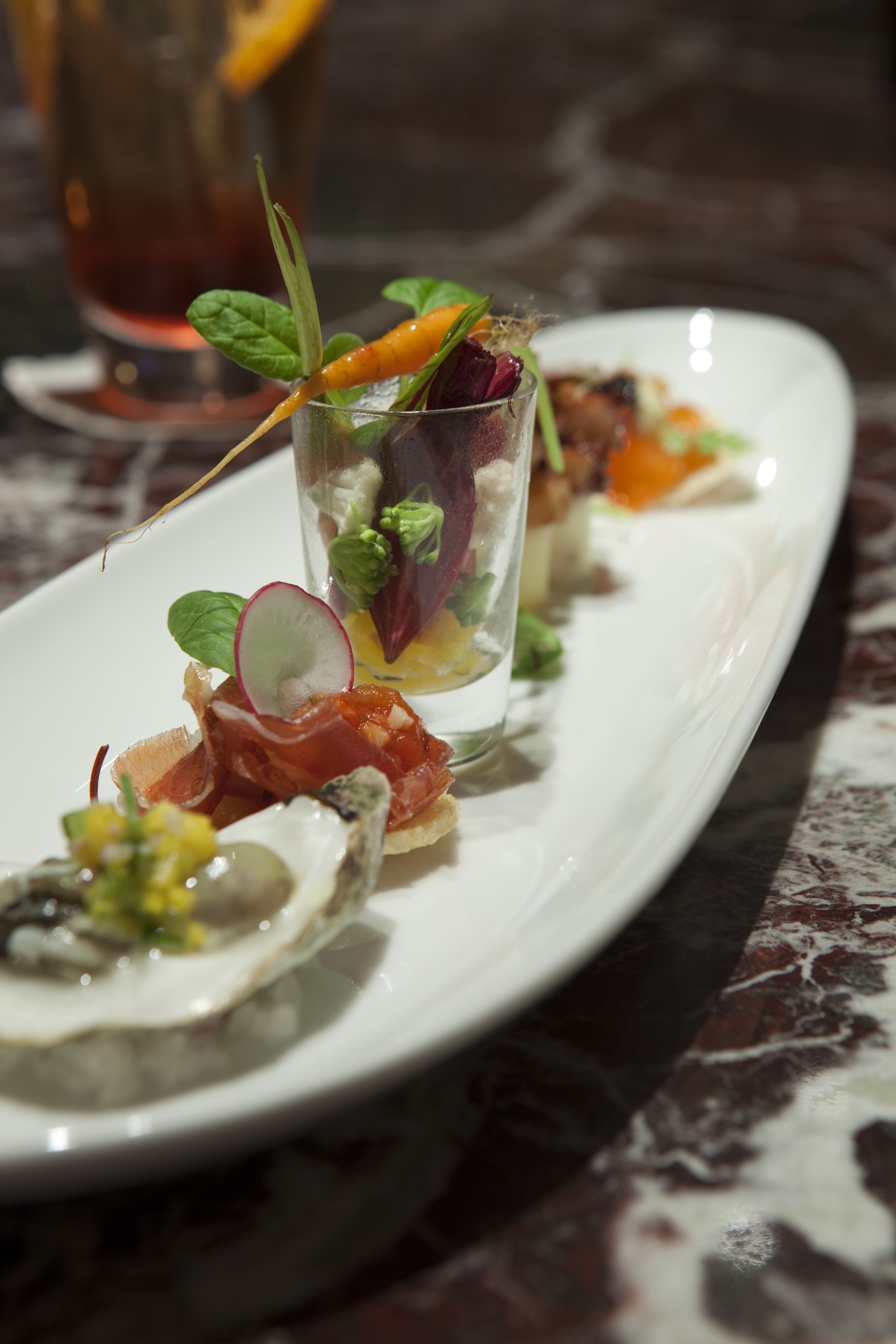 A white plate with various appetizers including oysters, a glass with a garnish, and a glass of red beverage.