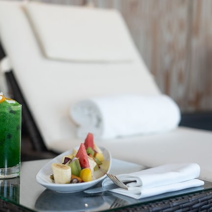 A glass of green beverage with a straw and a plate of fruit salad on a table in a room with a white chair and a wicker table.