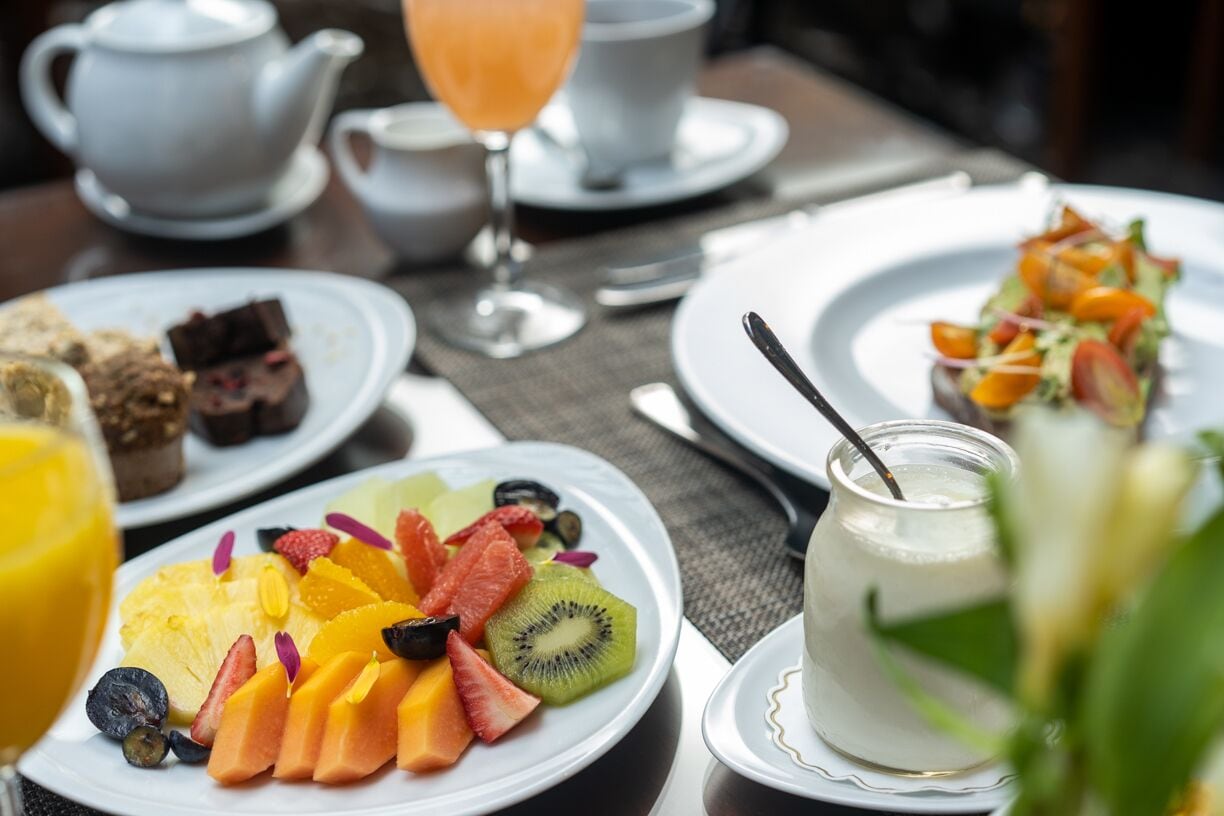 Una mesa de desayuno con platos de fruta, un vaso de jugo de naranja y una tetera.