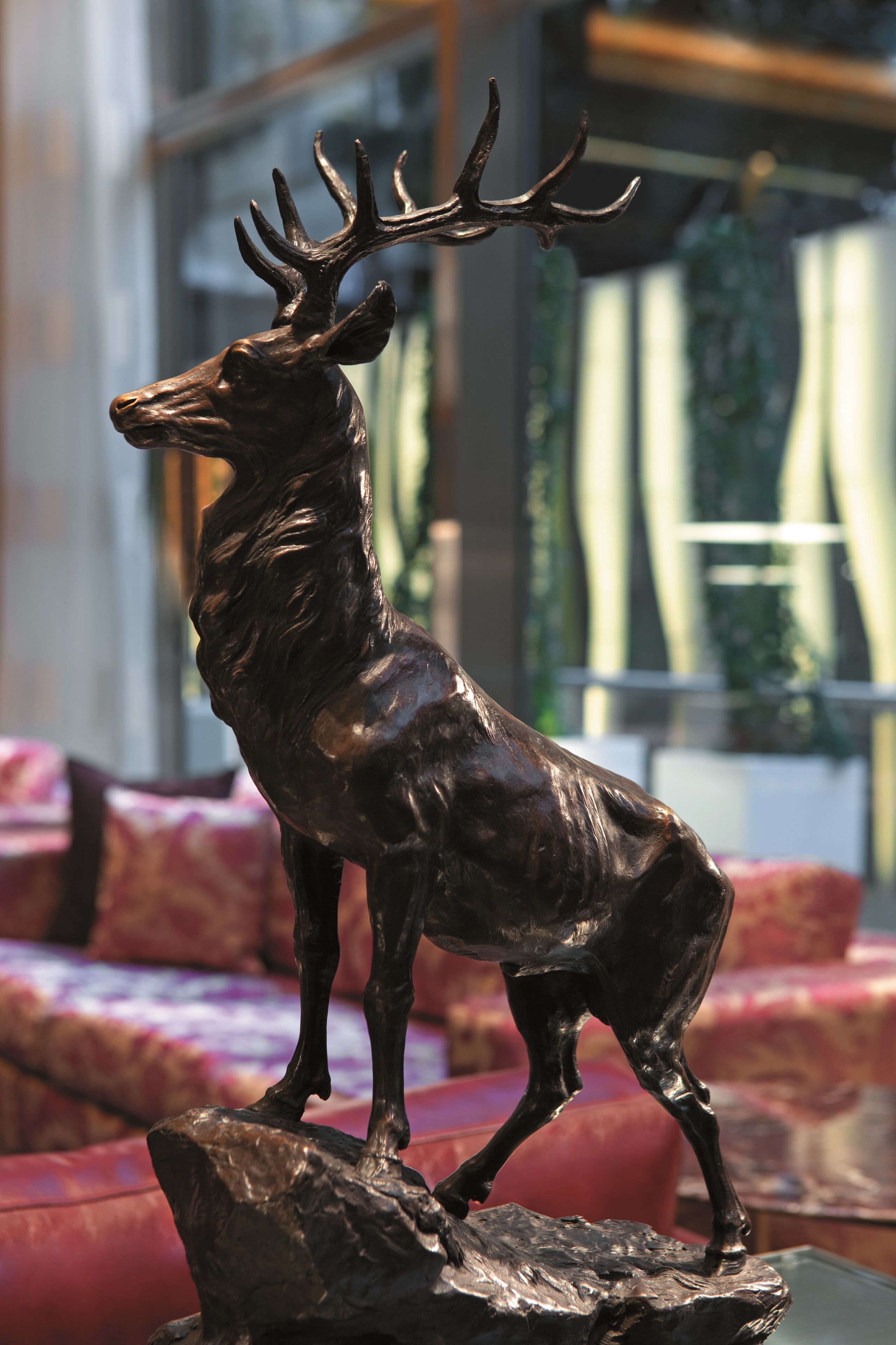 Una escultura de bronce de un ciervo sobre un pedestal en una habitación con un sofá rojo y una ventana con cortinas.