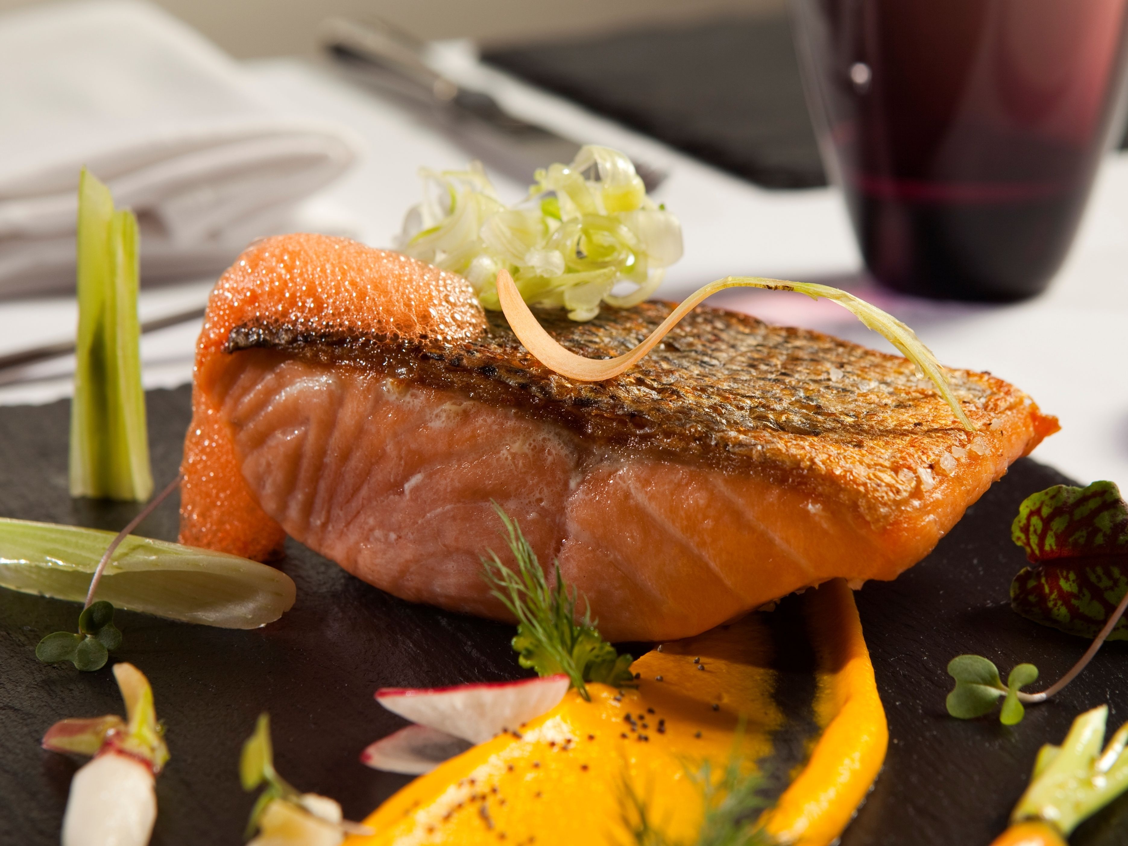 Een zalmfilet met knapperige huid, gegarneerd met kruiden en groenten, geserveerd op een zwart bord.