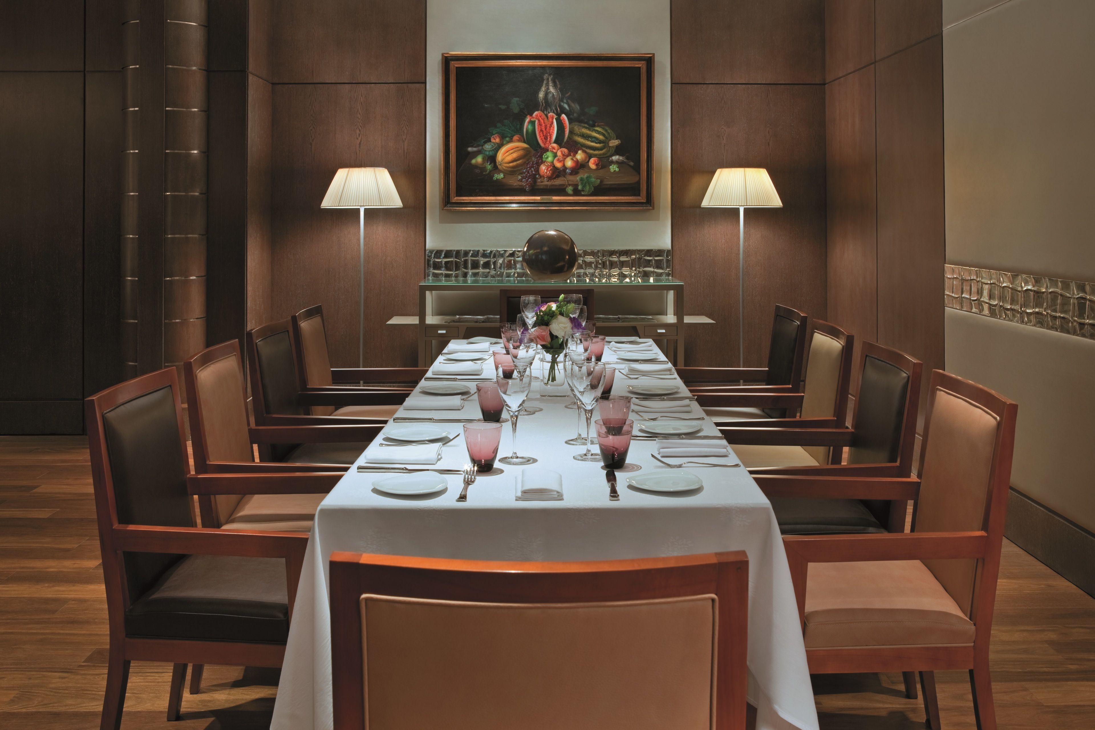 A formal dining room with a long table set for a meal, flanked by chairs, with a painting above and lamps on either side.