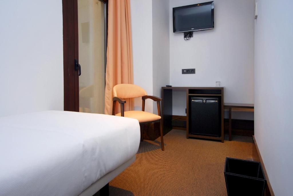 A hotel room with a bed, a chair, a desk, a television, and a trash bin.