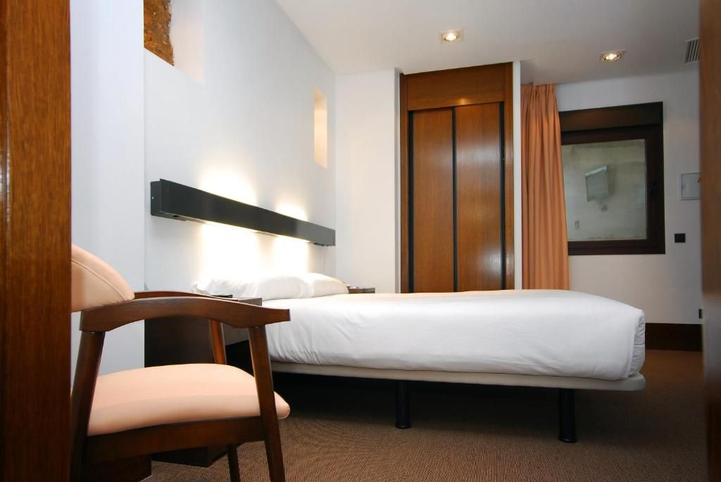 Una habitación de hotel con una cama, una silla, una mesita de noche y una lámpara fijada a la pared.