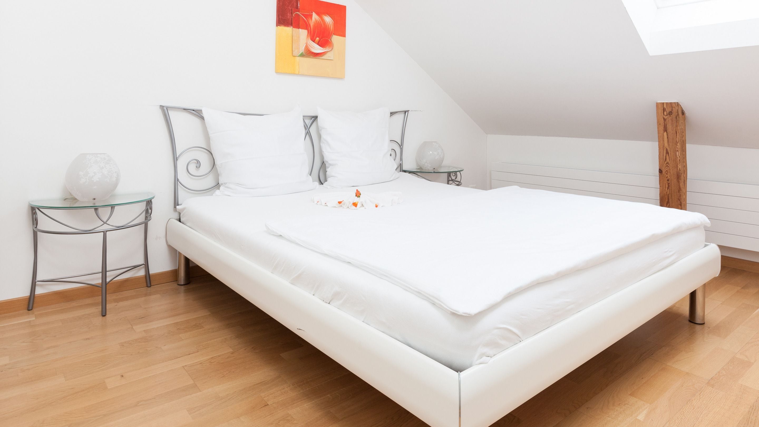 Una cama bien hecha en una habitación con techo inclinado, una mesita de noche pequeña con una lámpara, y una obra de arte colgada en la pared.
