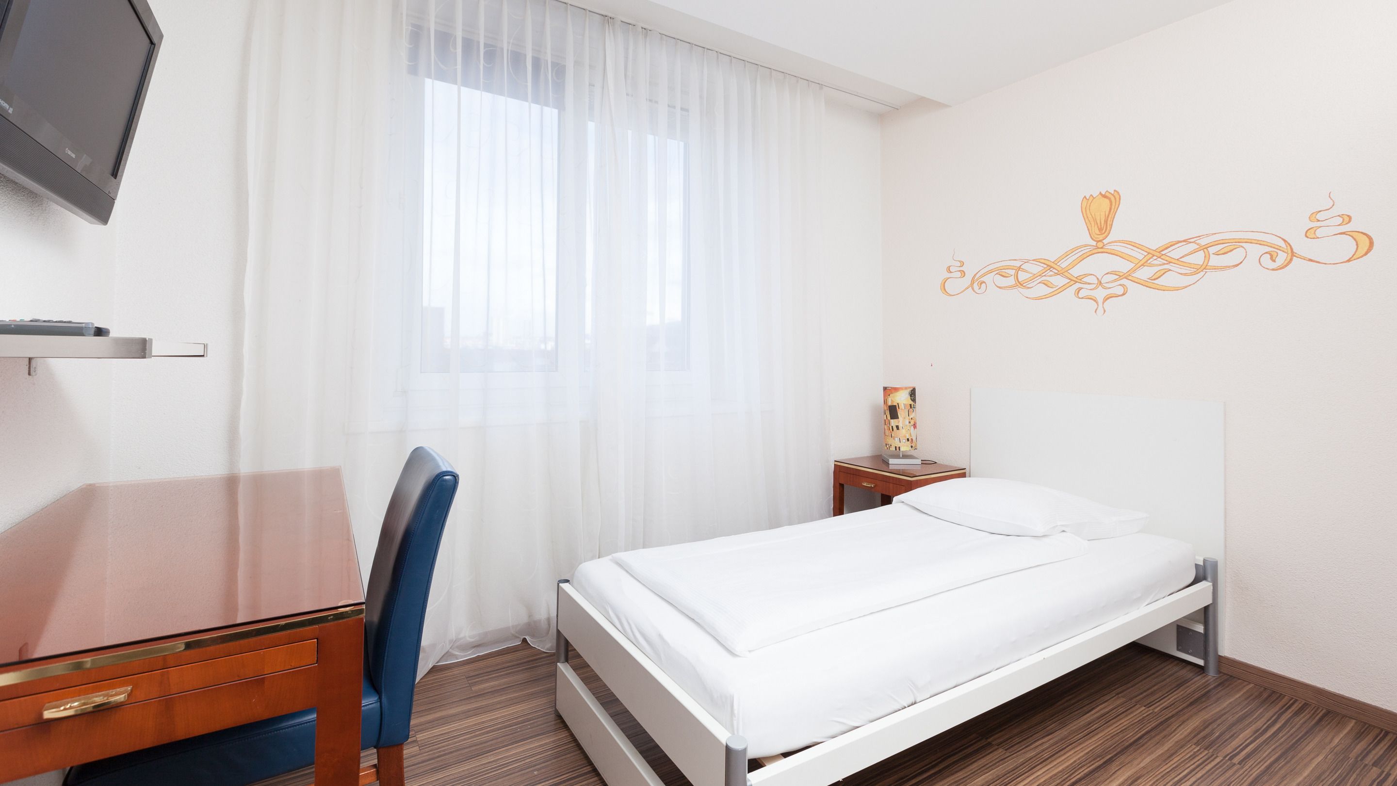 Habitación de hotel con cama, escritorio, silla, televisor y mesita de noche con lámpara.
