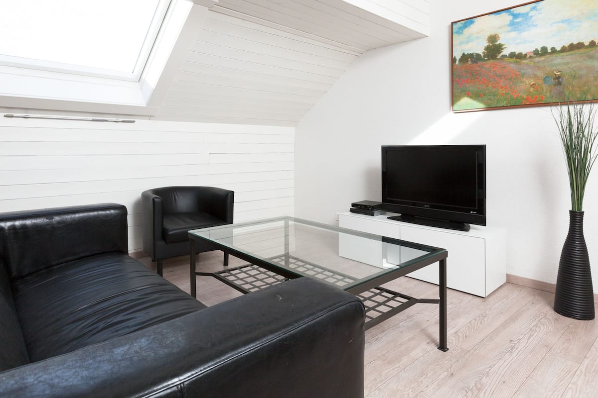 A modern living room with a black leather sofa, a glass coffee table, a black leather armchair, a flat-screen TV on a white cabinet, and a painting on the wall.