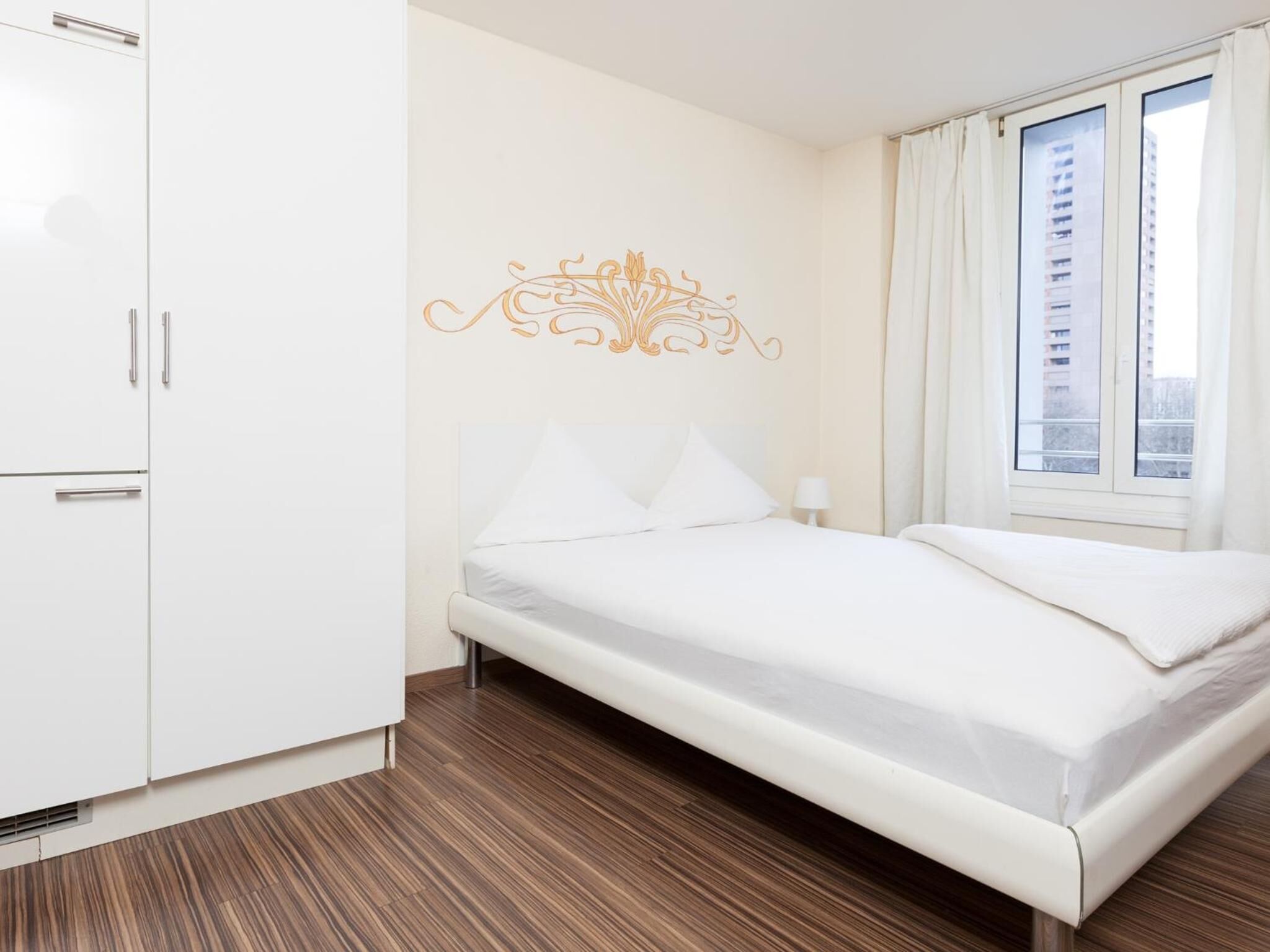 Ein modernes Hotelzimmer mit Holzboden, einem weißen Bett und einem Fenster mit Vorhängen.