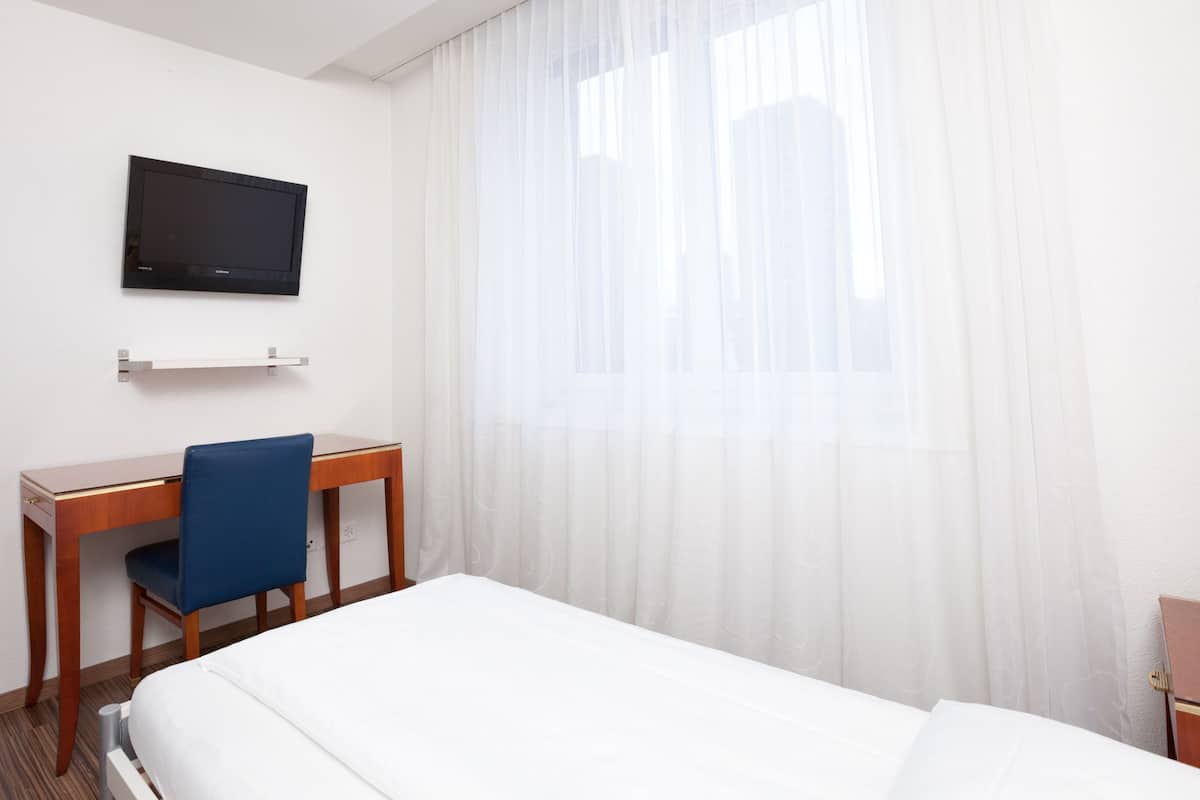 A hotel room with a bed, a desk, a chair, a television mounted on the wall, and a window with sheer curtains.
