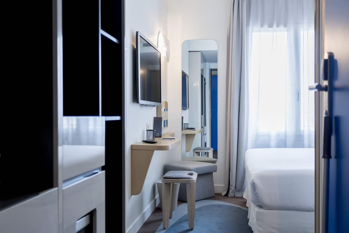 Habitación de hotel con una cama, un escritorio con computadora, un espejo y una ventana con cortinas.