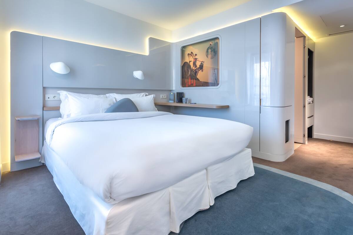 Una habitación de hotel moderna con una cama grande, una mesita de noche, un armario empotrado y una obra de arte colgada en la pared.