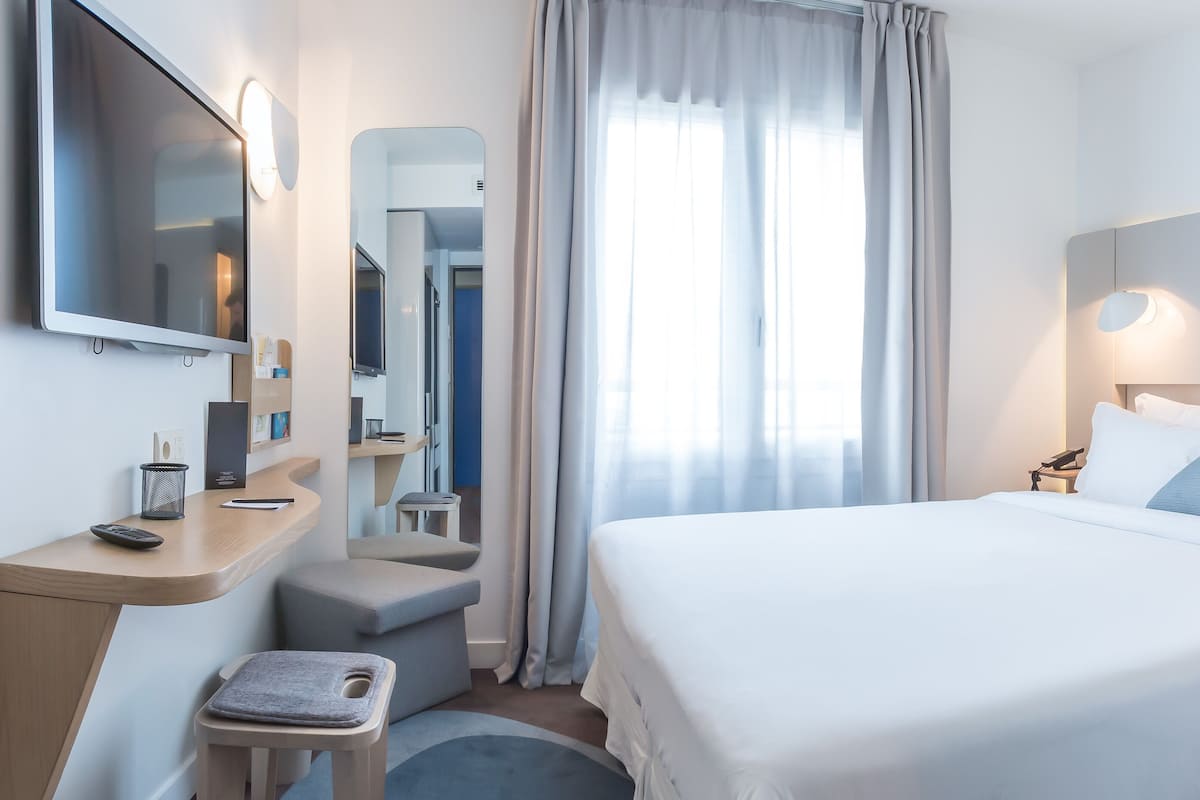 Una habitación de hotel moderna con una cama, un escritorio, una silla, un espejo y una ventana con cortinas.