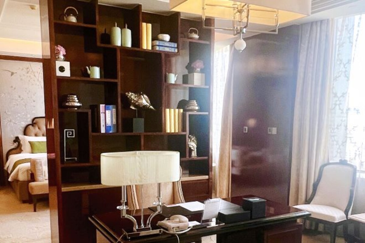 Una habitación de hotel con una amplia estantería de madera, un escritorio con lámpara, una silla y una cama con mesita de noche.