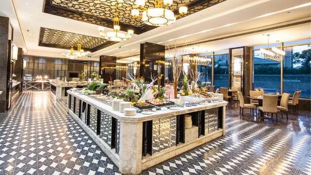 Un moderno vestíbulo de hotel con piso a cuadros, una zona de bar central con diversas opciones de comida y un gran ventanal con vistas al exterior.