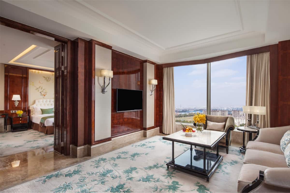 A hotel room with a large window, a flat-screen TV, a sofa, a coffee table, and a bed with a headboard.