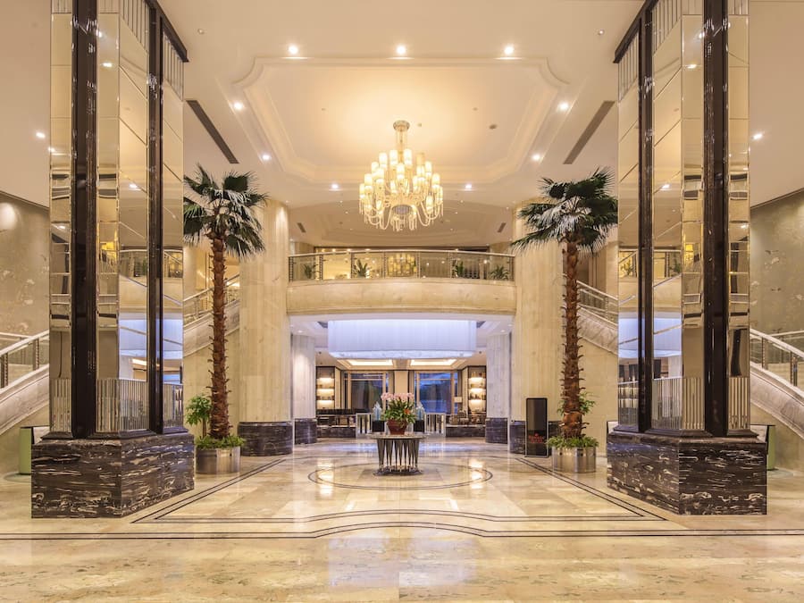 A spacious lobby with a grand chandelier, marble flooring, and a central reception desk.