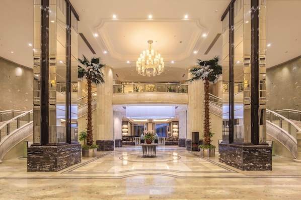 A spacious lobby with a grand chandelier, marble flooring, and a central reception desk.