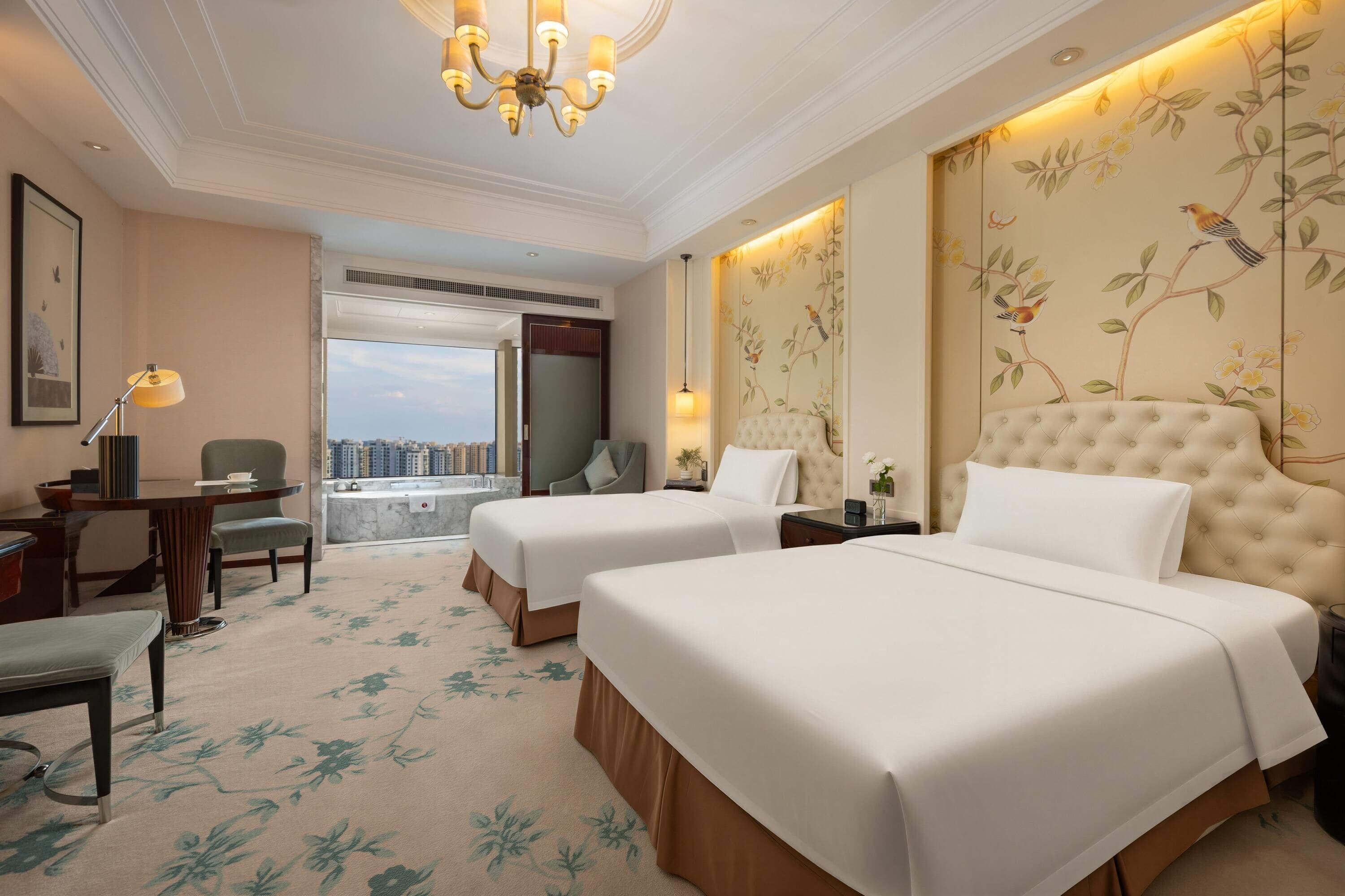 A hotel room with two beds, a desk, a chair, a view of the city, and a chandelier.