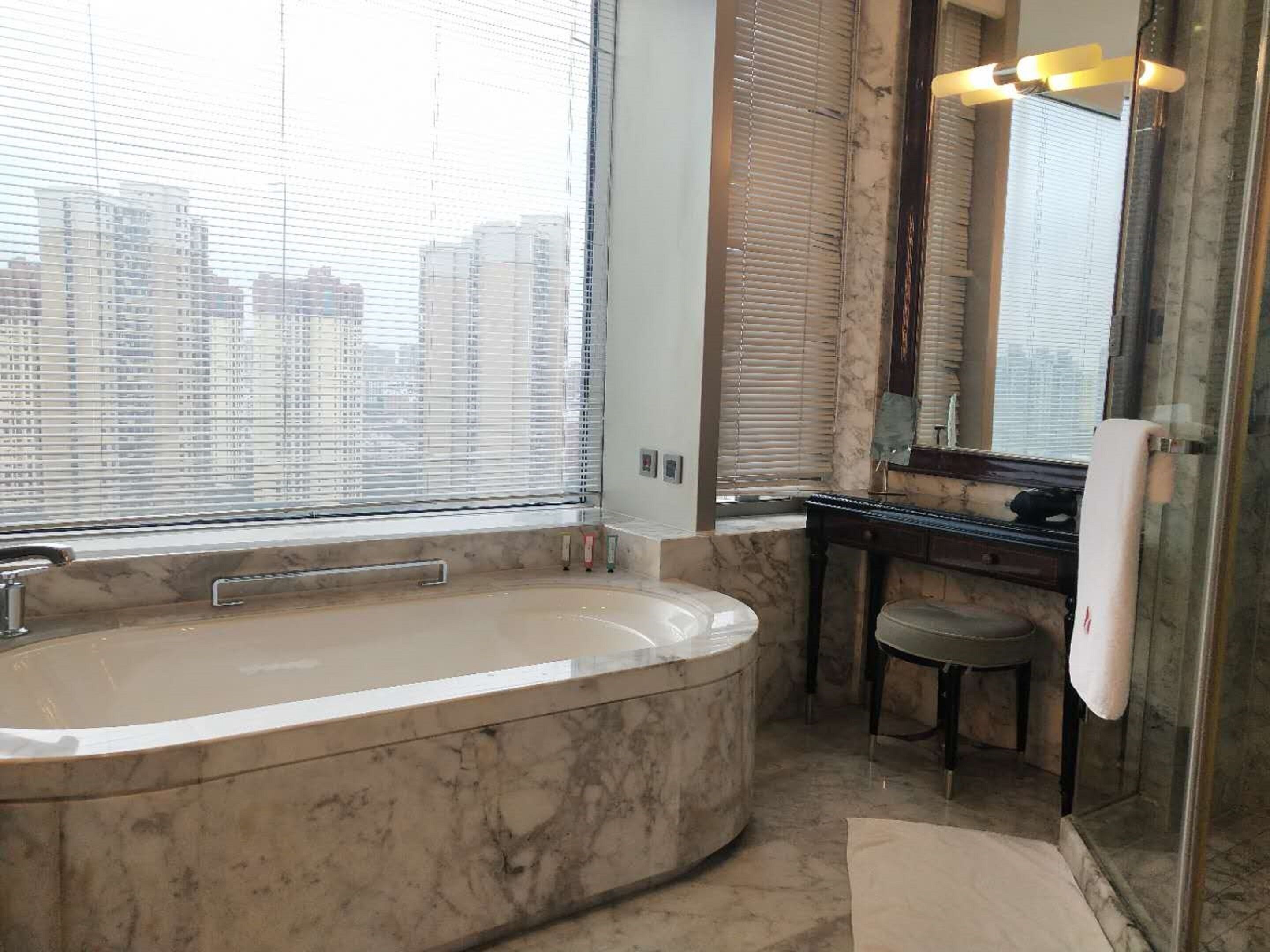 A modern bathroom with a large bathtub, a vanity with a mirror, and a city view through the windows.