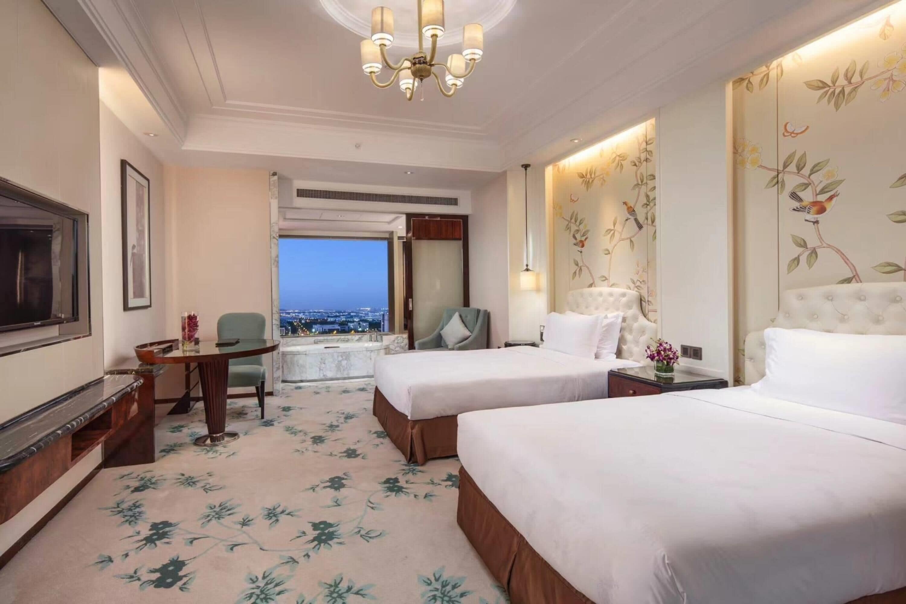 Ein Hotelzimmer mit zwei Betten, einem Schreibtisch, einem Sessel, einem Fernseher und Blick auf die nächtliche Stadt.
