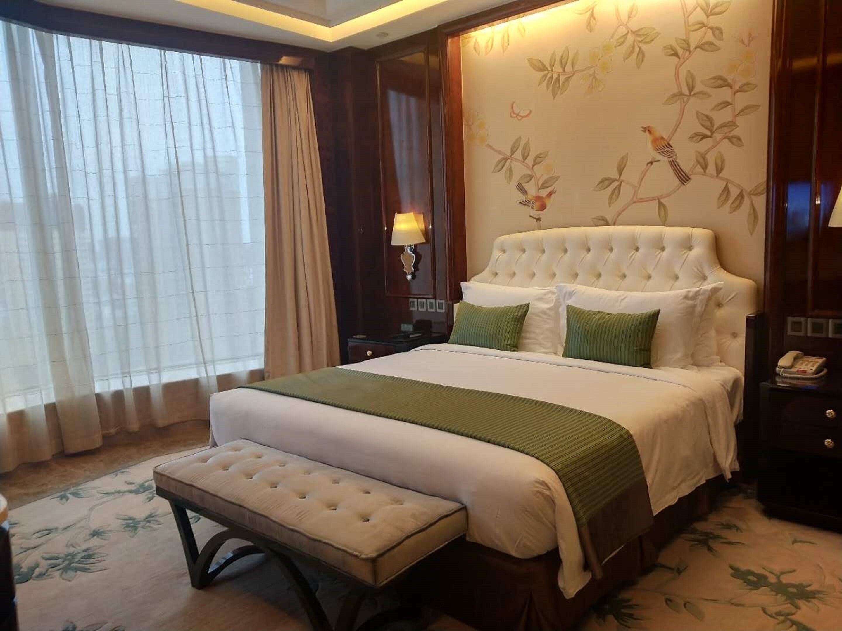 A hotel room with a large bed, a bench, a nightstand, and a wall with a floral pattern.